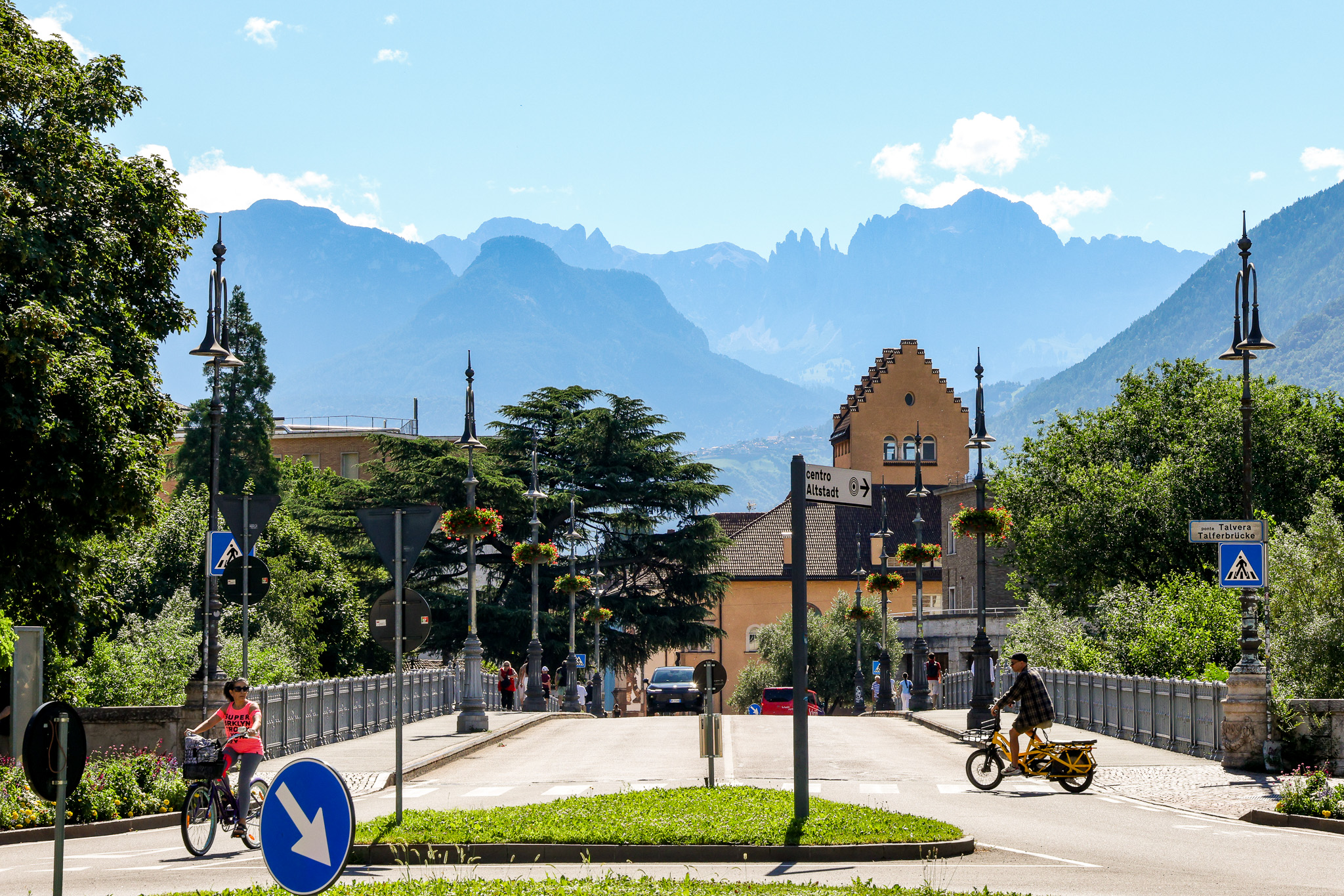 A wide photographic view of Bolzano’s historic city center, focusing on elegant stone facades around Waltherplatz. Detailed pastel-colored buildings with carved window frames and flower boxes line a clean cobbled square, with the Gothic spire of the Bolzano Cathedral rising sharply in the background. Soft late-afternoon sunlight bathes the scene, casting long, gentle shadows and bringing out the warm tones of the stone. The mood is welcoming and professional, emphasizing discovery and history. Shot at eye level with sharp focus throughout, using a balanced composition that leaves space in the sky area for potential website text overlay. Photographic realism, crisp and clear, with no people visible, just an inviting, orderly urban landscape.